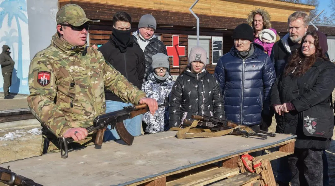 Інструктор із шевроном підрозділу показує цивільним автомат АК; на столі ще кілька одиниць зброї, довкола стоять…