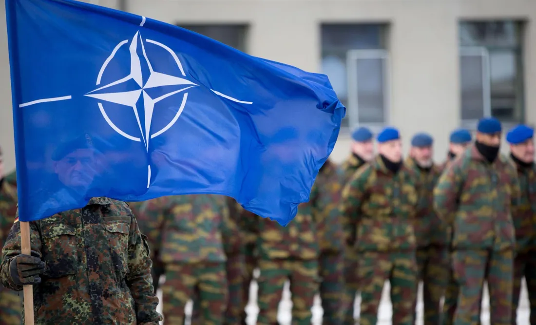 Синій прапор НАТО з компасною розою на передньому плані, позаду розмита шеренга військових у камуфляжі й блакитних…