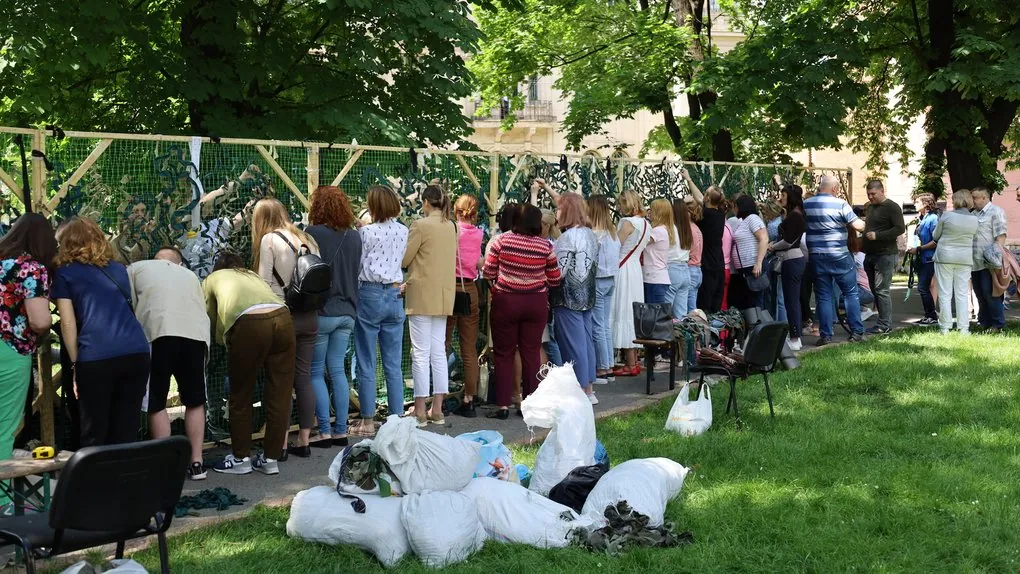 Люди різного віку стоять уздовж натягнутої сітки й вплітають у неї стрічки — виготовлення маскувальних сіток для фронту.