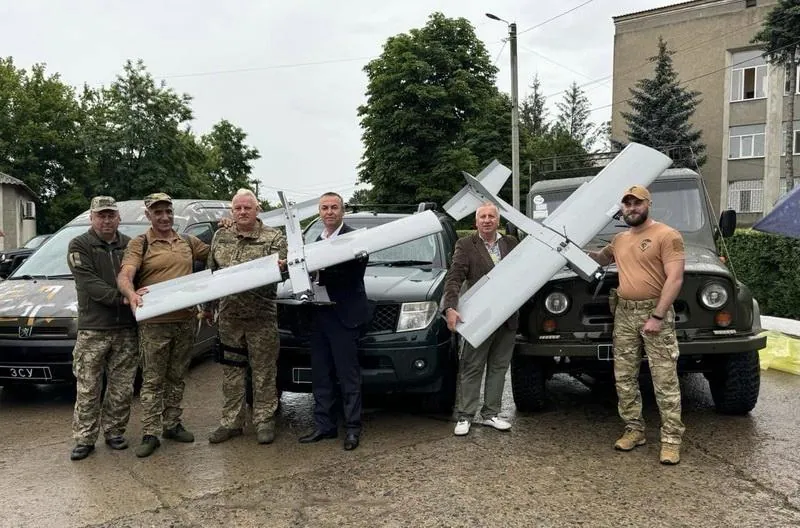 Група військових і цивільних тримають два розвідувальні БпЛА літакового типу на тлі переданих позашляховиків із написом…