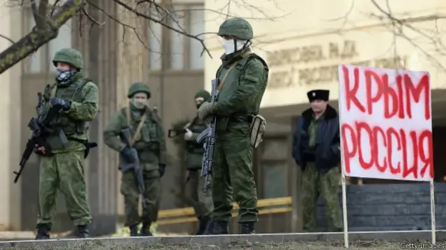 Озброєні військові у зеленій формі без розпізнавальних знаків («зелені чоловічки») біля адмінбудівлі; поруч плакат…