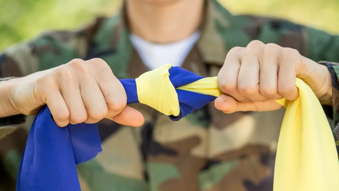 Руки, стиснуті в кулаки, тримають скручені синю і жовту тканини на тлі камуфляжної форми — національна єдність…