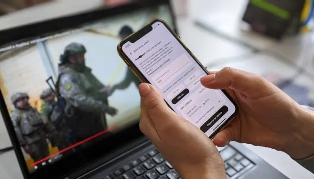 Руки тримають смартфон із формою онлайн-донату; на екрані ноутбука позаду — відео з військовими. Збір коштів на армію.