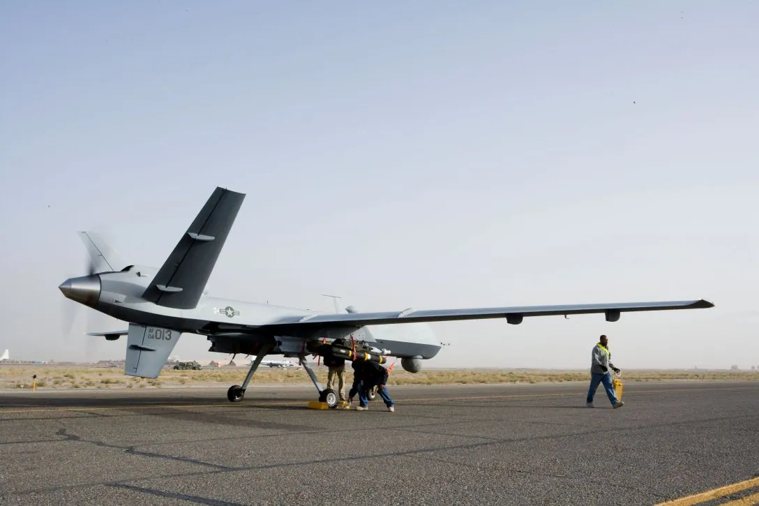 Великий ударно-розвідувальний БпЛА типу MQ-9 Reaper на злітній смузі, поруч техніки; під крилами підвіски.