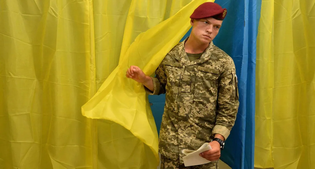 Військовослужбовець у піксельній формі з червоним береєм стоїть біля виборчої кабіни, задрапованої прапором України; у…