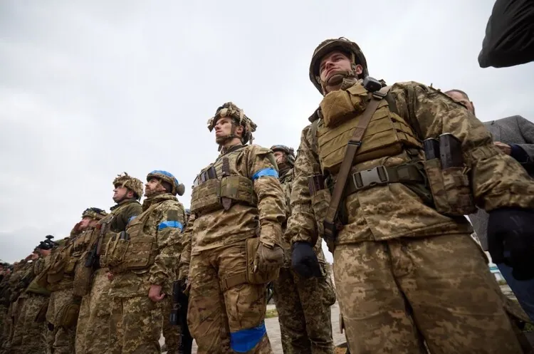 Шеренга військових ЗСУ у бронежилетах і касках, знято знизу; на руках синьо-жовті розпізнавальні пов'язки.