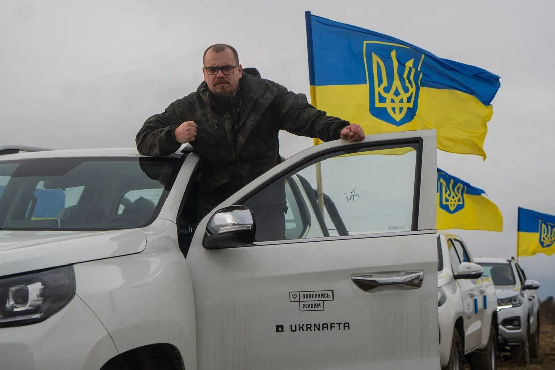 Чоловік біля білого пікапа з наліпкою «Повернись живим» і логотипом Ukrnafta; поряд прапори України з тризубом, у ряду…
