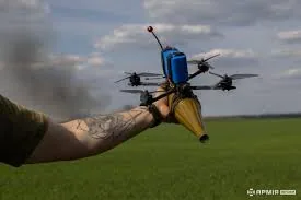 Рука в рукавичці тримає FPV-дрон із підвішеним боєприпасом у полі; на задньому плані дим — бойове застосування FPV.
