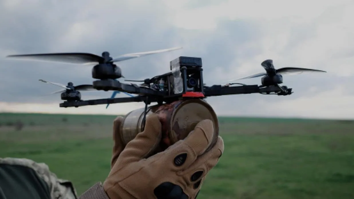 Рука в тактичній рукавичці тримає FPV-дрон із закріпленим кумулятивним боєприпасом; за спиною зелене поле — техніка…