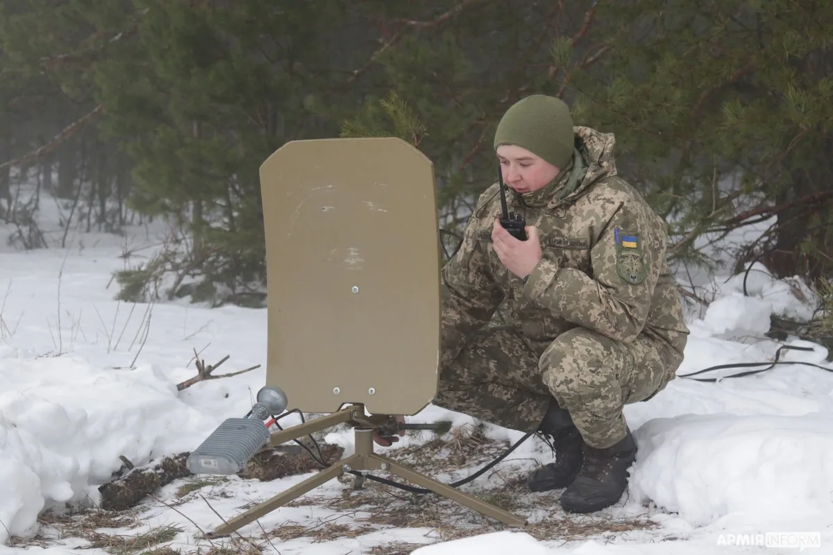 Зв'язківиця в зимовому полі говорить у радіостанцію, поруч на триногі розгорнута плоска супутникова/спрямована антена…
