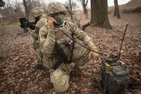 Боєць у лісі на колінах працює з переносною радіостанцією у рюкзаку, поруч другий прикриває — достовірність і стійкість…