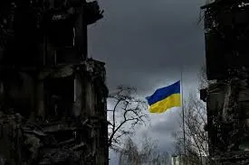 Прапор України на флагштоку серед вигорілих зруйнованих будинків під темним небом — воля і стійкість громадян.